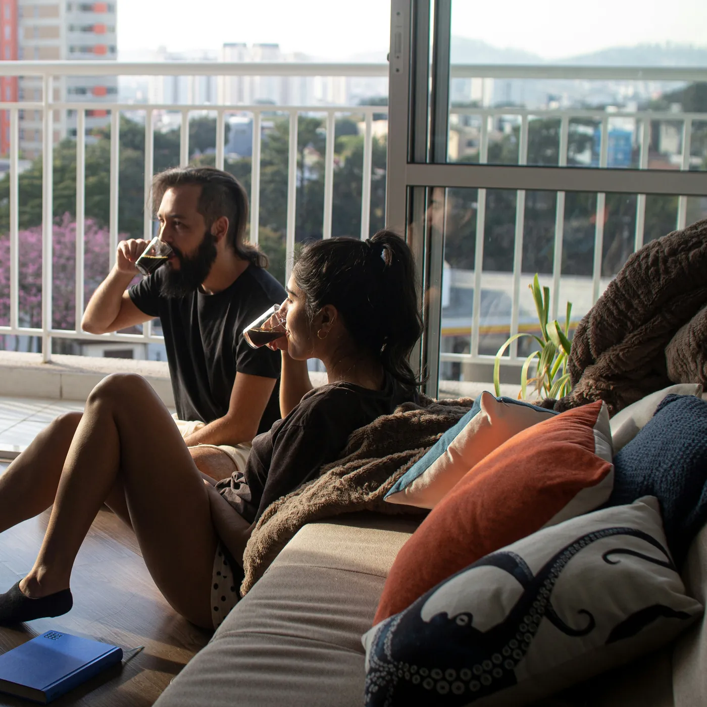 Two people sitting on the floor by an open balcony, drinking coffee and looking out over the city together.